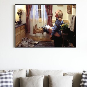 May include: A framed photograph of a man sitting on a toilet with his legs raised in the air. He is wearing a blue shirt and jeans. The bathroom is decorated with a floral arrangement and a window with white curtains.