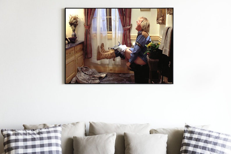 May include: A framed photograph of a man sitting on a toilet with his legs raised in the air. He is wearing a blue shirt and jeans. The bathroom is decorated with a floral arrangement and a window with white curtains.