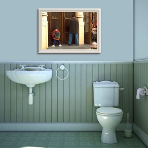 Pode incluir: Uma fotografia emoldurada de um homem e um menino em pé na frente de um prédio com as palavras "Felix" e "Toiletten" escritas nas portas. A fotografia está pendurada em uma parede azul clara em um banheiro com uma pia de porcelana branca, um vaso sanitário e um piso de ladrilhos.