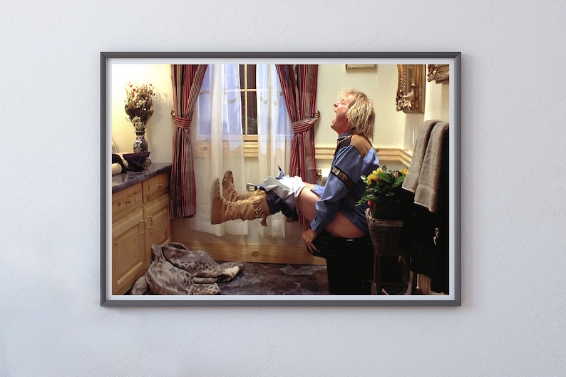 May include: A man in a blue shirt and brown boots sits on a toilet with his legs raised in the air. He is screaming with his mouth open. The bathroom is decorated with a floral arrangement, a framed picture, and a towel rack.