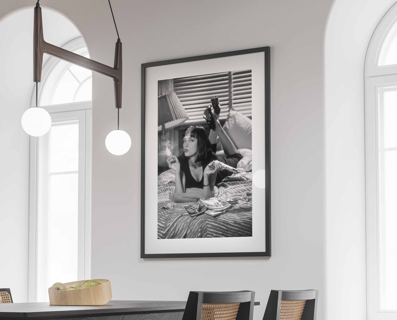Op de afbeelding: Zwart-witfoto van een vrouw die op een bed ligt, een sigaret rookt en een stapel geld vasthoudt. De vrouw draagt een zwart topje en heeft haar haar naar achteren getrokken. De foto is ingelijst in een zwart frame.