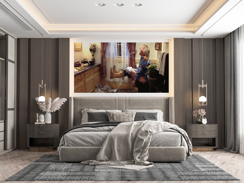 May include: A bedroom interior with a large framed print of a man sitting on a toilet. The print is hanging on a wall with a dark brown wood paneling. The bed is made with a gray duvet and a white throw blanket. There are two nightstands with lamps on either side of the bed.