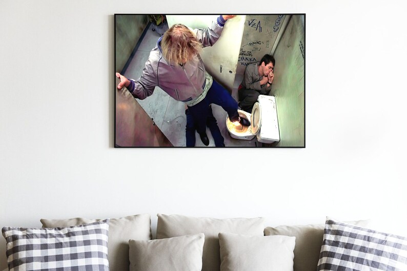 K&ouml;nnte beinhalten: Ein Schwarzwei&szlig;foto von zwei Personen in einem Badezimmer. Eine Person steht auf einer Toilette mit dem Fu&szlig; auf dem Kopf der anderen Person. Die Badezimmerw&auml;nde sind mit Graffiti bedeckt.