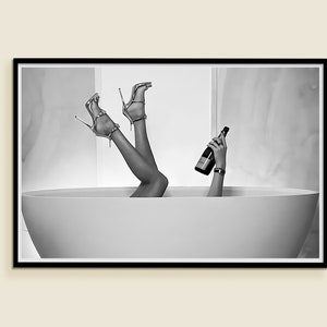 Op de afbeelding: Zwart-witfoto van benen en voeten die uit een wit bad steken. De persoon houdt een fles champagne vast.
