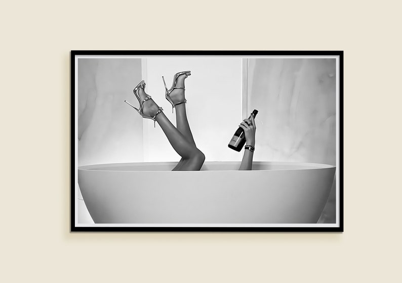 Op de afbeelding: Zwart-witfoto van benen en voeten die uit een wit bad steken. De persoon houdt een fles champagne vast.