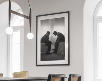 May include: Black and white photograph of two men sitting on a couch, looking down. The photo is framed in a black frame and hanging on a white wall.