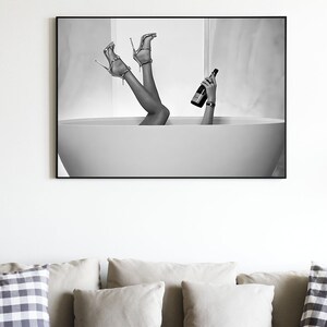 Op de afbeelding: Zwart-witfoto van benen en voeten die uit een badkuip steken, met een fles champagne in de hand.