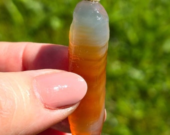 May include: A smooth, polished agate stone with a light gray and white top and a deep orange bottom. The stone is shaped like a long, thin cylinder.