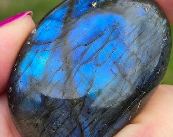 May include: Oval-shaped labradorite stone with a striking display of blue iridescence. The polished surface reflects light, showcasing a spectrum of blues and grays. The stone is held in a hand, highlighting its natural beauty and unique patterns.