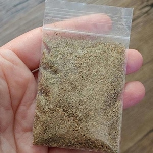 May include: A small, clear plastic bag filled with a finely ground, light brown substance. The bag is sealed at the top and held in a person's hand, with the background showing a wooden surface.