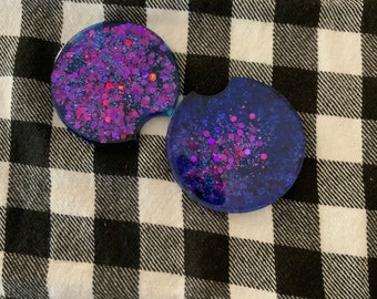 Puede incluir: Dos posavasos redondos para coche con diseño brillante. Uno es de color púrpura oscuro con purpurina rosa y roja, y el otro es azul oscuro con purpurina rosa. Ambos tienen una pequeña muesca para facilitar la extracción.