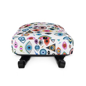 Puede incluir: Una mochila blanca con un patr&oacute;n de ojos coloridos. Los ojos son de varios colores, incluyendo azul, verde, rojo y amarillo. La mochila tiene correas ajustables y un cierre de cremallera.