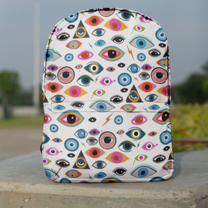 Puede incluir: Una mochila blanca con un patr&oacute;n de ojos coloridos. Los ojos son de varios colores, incluyendo azul, verde, rosa y naranja. Los ojos est&aacute;n rodeados de un fondo blanco con rayos y tri&aacute;ngulos.