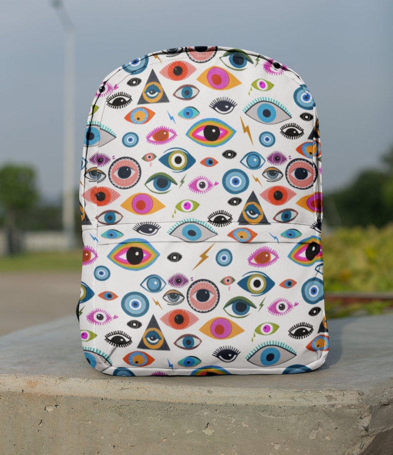 Puede incluir: Una mochila blanca con un patr&oacute;n de ojos coloridos. Los ojos son de varios colores, incluyendo azul, verde, rosa y naranja. Los ojos est&aacute;n rodeados de un fondo blanco con rayos y tri&aacute;ngulos.