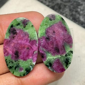 May include: Two oval-shaped ruby zoisite gemstones. The gemstones feature a vibrant mix of colors, with a dominant magenta hue interspersed with green and black patches. The stones are polished and ready for jewelry making.