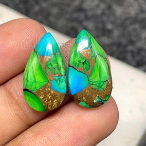 May include: Two teardrop-shaped gemstones with vibrant colors. The stones feature a mix of bright green, turquoise blue, and gold-colored matrix patterns. The gemstones are held in a person's hand.