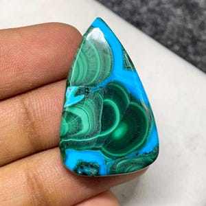 May include: A polished gemstone with a teardrop shape. The stone features a vibrant mix of teal and green hues, with swirling patterns and a glossy finish. The gemstone is held in a hand, showcasing its natural beauty.