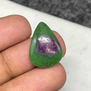 Könnte beinhalten: Ein tropfenförmiger Edelstein mit grüner Außenseite und einem zentralen, unregelmäßigen violetten Einschluss. Der Stein wird in einer Hand gehalten, wodurch seine polierte Oberfläche und die natürlichen Farbvariationen zur Geltung kommen. Die Form des Edelsteins erinnert an einen Anhänger oder Cabochon.