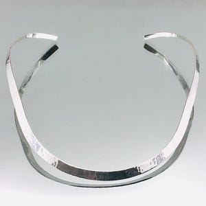以下が含まれることがあります： ハンマー加工が施されたシルバーカラーのチョーカーネックレス。 ネックレスは、首に巻き付くようにカーブしたオープンデザインです。 金属は光を反射し、ハイライトと影を作り出します。 全体的なスタイルはモダンでミニマリストです。