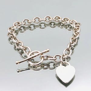 Op de afbeelding: Een zilveren kettingarmband met een hartvormige hanger en een schuifsluiting.