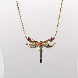 May include: A gold-tone necklace featuring a dragonfly pendant with openwork wings. The dragonfly is decorated with inlaid teardrop-shaped pink stones, small orange and blue accents, and a central oval orange stone on a delicate gold chain.