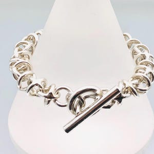 Può includere: Un braccialetto color argento con chiusura a toggle. Il braccialetto presenta un design a catena con maglie a forma di stella. La chiusura a toggle è una barra che si inserisce in un anello circolare. Il braccialetto è esposto su un supporto bianco.