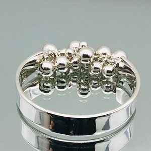 Könnte beinhalten: Ein silberner Ring mit einem Cluster aus kleinen, runden Perlen. Die Perlen sind in einem zufälligen Muster angeordnet und durch ein dünnes silbernes Band verbunden. Der Ring ist breit und hat eine glatte, polierte Oberfläche.