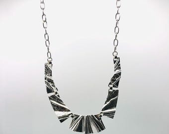 Puede incluir: Un collar plateado con un diseño geométrico. El collar presenta una cadena y un colgante compuesto por varias piezas rectangulares y triangulares. El colgante tiene un aspecto texturizado y veteado con patrones en blanco y negro.