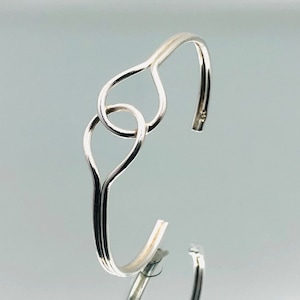 Könnte beinhalten: Ein silbernes Armband mit einem Doppelherz-Design. Das Armband besteht aus einem einzigen Stück Draht, das zu zwei ineinander verschlungenen Herzformen gebogen ist.