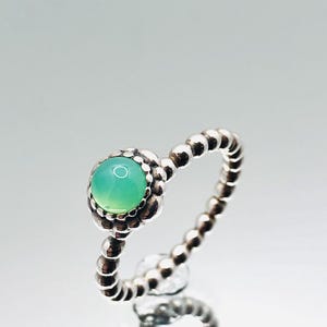 May include: A silver ring with a round, light green gemstone at the center. The band features a beaded design. The gemstone is surrounded by a floral-like setting.