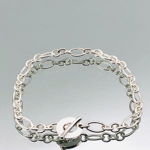 May include: A silver bracelet featuring an oval link chain design. The bracelet has a toggle clasp closure. The jewelry is displayed on a reflective surface.