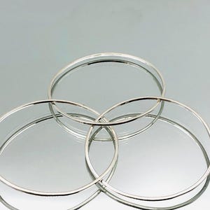 May include: Three silver bangle bracelets stacked on top of each other. The bracelets are all the same width and have a smooth, polished finish.