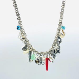 May include: A silver chain necklace with multiple charms, including a red coral charm, a turquoise charm, a feather charm, and a shell charm.