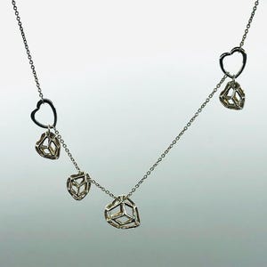 Può includere: Una collana d'argento con quattro ciondoli. I ciondoli sono a forma di cuore e forme geometriche. La collana è su uno sfondo bianco.