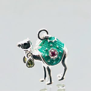 May include: A silver charm in the shape of a camel with a green and red patterned saddle. The camel is decorated with green and clear gemstones.