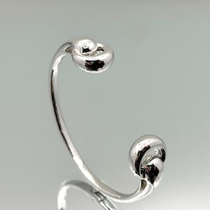 May include: A polished silver-tone open cuff bracelet featuring two rounded, knot-like terminals at each end. The metal surface is smooth and reflective, creating a continuous curved band that remains open at the front for easy wear.