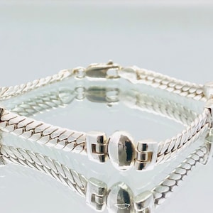 May include: A silver bracelet with a woven chain design and a round, silver charm. The bracelet has a clasp closure.