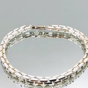Könnte beinhalten: Ein silbernes Armband mit geometrischem Design. Das Armband besteht aus rechteckigen Gliedern, die miteinander verbunden sind. Der Verschluss ist sichtbar und scheint sicher zu sein. Das Armband wird auf einer reflektierenden Oberfläche präsentiert.