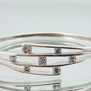 Könnte beinhalten: Ein silbernes Armreif-Armband mit einem zarten Design mit mehreren Bändern und klaren, funkelnden Steinen.