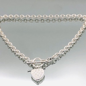 Op de afbeelding: Zilverkleurige ketting met een hartvormige hanger. De ketting heeft een dubbel kettingontwerp en een tuimelsluiting. De hartvormige hanger is getextureerd en reflecteert licht.