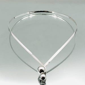 May include: A silver necklace with a minimalist design. The necklace has a thin, curved band forming a V-shape, joined by small, round beads. The reflective surface enhances the jewelry's elegant appearance. The necklace is likely made of sterling silver.