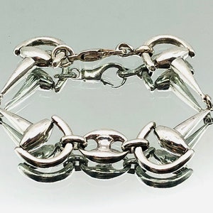Puede incluir: Un brazalete de plata con un diseño de bocado de caballo. El brazalete está hecho de eslabones entrelazados que tienen forma de bocado de caballo.