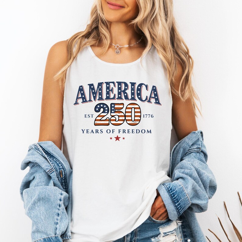 May include: White tank top with the text "AMERICA 250 YEARS OF FREEDOM" in blue, red, and white. The design includes stars and the year 1776. The tank top is worn with a denim jacket.
