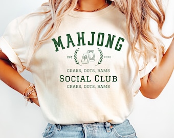 Puede incluir: Una camiseta de color crema con texto en verde oscuro que dice MAHJONG SOCIAL CLUB, EST 2026 y CRAKS, DOTS, BAMS. El diseño incluye un gráfico de dos fichas de mahjong rodeadas por una corona de laurel, impreso en la parte frontal.