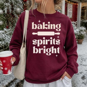 K&ouml;nnte beinhalten: Ein kastanienbraunes Sweatshirt mit Rundhalsausschnitt und dem cremefarbenen Schriftzug "baking spirits bright", einer Nudelholz-Grafik und kleinen Sternen. Das Kleidungsst&uuml;ck hat lange &Auml;rmel und einen gerippten Kragen, kombiniert mit einer beigen Stofftasche.