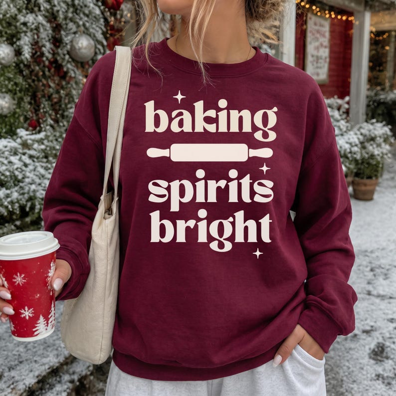 K&ouml;nnte beinhalten: Ein kastanienbraunes Sweatshirt mit Rundhalsausschnitt und dem cremefarbenen Schriftzug "baking spirits bright", einer Nudelholz-Grafik und kleinen Sternen. Das Kleidungsst&uuml;ck hat lange &Auml;rmel und einen gerippten Kragen, kombiniert mit einer beigen Stofftasche.