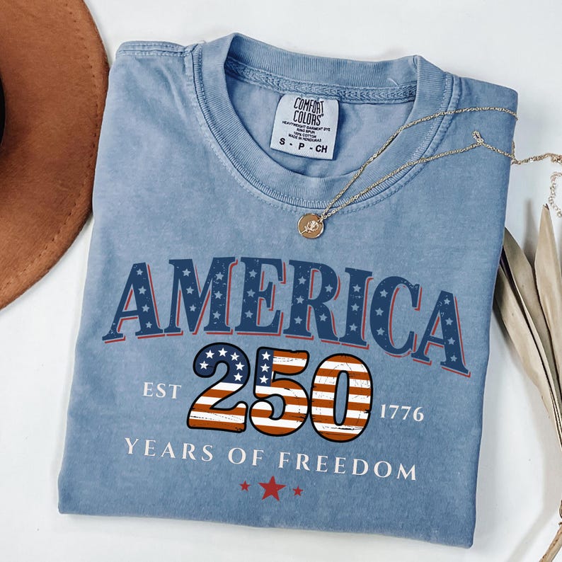 May include: A light blue t-shirt with the word "AMERICA" in navy blue and red lettering, with stars. The number "250" is in the center, with the American flag design. The text "EST 1776" and "YEARS OF FREEDOM" are also visible.