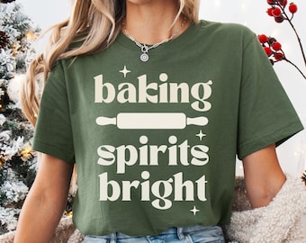 May include: An olive green short-sleeve t-shirt featuring the cream-coloured text "baking spirits bright" with a graphic of a rolling pin in the centre and small four-pointed star accents printed on the front of the garment.