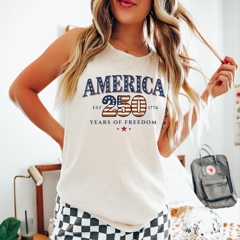 May include: Cream-colored tank top with the word "AMERICA" in navy blue, and "250" in red and blue, with "EST 1776" and "YEARS OF FREEDOM" below. The design features stars and stripes, celebrating the nation's history.
