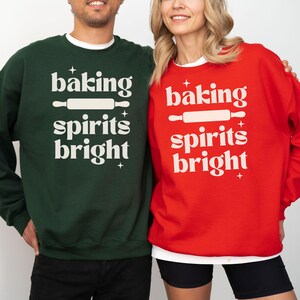 K&ouml;nnte beinhalten: Ein dunkelgr&uuml;nes Sweatshirt und ein leuchtend rotes Sweatshirt, beide mit dem wei&szlig;en Schriftzug "baking spirits bright" und einer Nudelholz-Grafik. Das Design enth&auml;lt kleine vierzackige Sterne oberhalb und unterhalb des Textes.